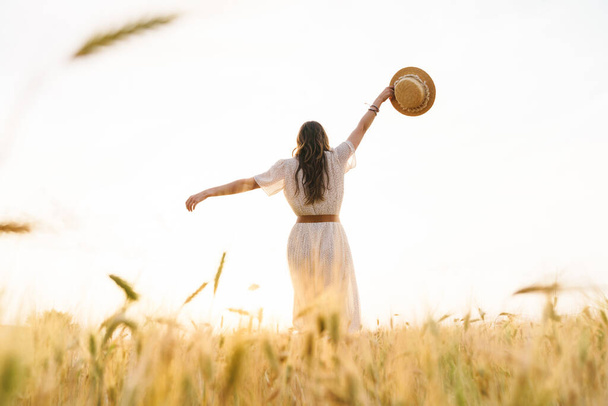Photo from back of cute brunette woman with straw hat dancing on wheat field at summer day - Foto, Imagem