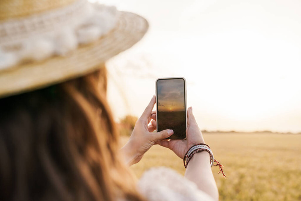 Photo of young woman in straw hat talking photo on cellphone while walking on wheat field at summer day - Foto, Imagem