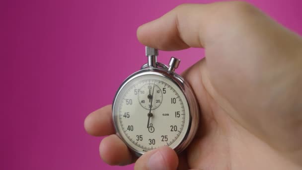 Mão masculina segurando cronômetro analógico em fundo rosa. O tempo começa com o velho cronômetro homem pressiona botão iniciar no conceito de esporte. Conceito de gestão do tempo
. - Filmagem, Vídeo