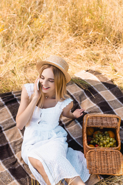 high angle view of blonde woman in summer outfit talking on smartphone near wicker basket with fruits - Foto, immagini