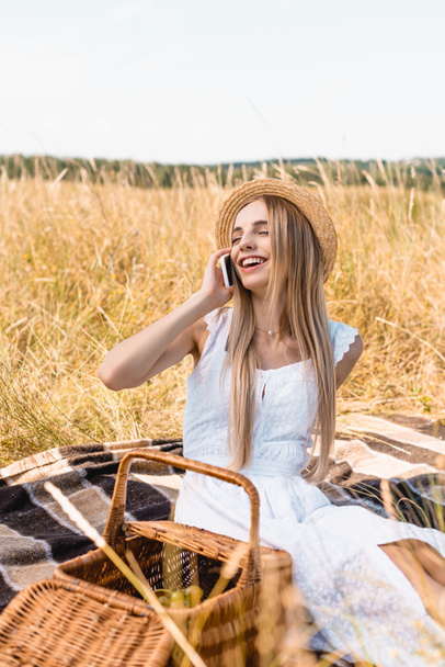 Selektiver Fokus einer aufgeregten Frau in weißem Kleid und Strohhut, die in Korbnähe auf dem Smartphone spricht  - Foto, Bild