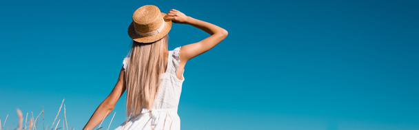 back view of blonde woman in white dress touching straw hat against blue sky, panoramic concept - Foto, Imagen