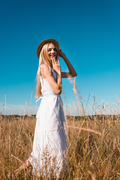 enfoque selectivo de la mujer sensual en vestido blanco tocando sombrero de paja y sosteniendo la mano cerca de la barbilla mientras mira a la cámara - Foto, imagen