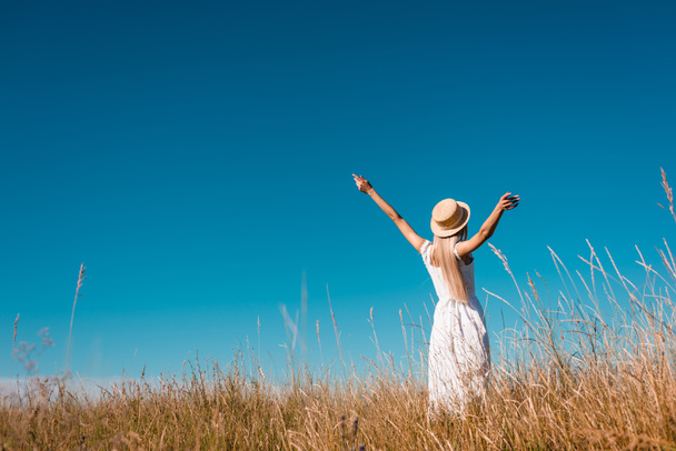 Rückansicht einer Frau in weißem Kleid und Strohhut, die mit ausgestreckten Händen im Grasfeld vor blauem Himmel steht - Foto, Bild