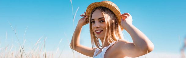 selective focus of young blonde woman touching straw hat and looking at camera against blue sky, panoramic concept - Foto, Imagem