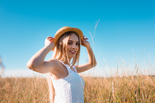 Hasır şapkaya dokunan ve çimen tarlasında kameraya bakan şehvetli sarışın kadının seçici odağı. - Fotoğraf, Görsel