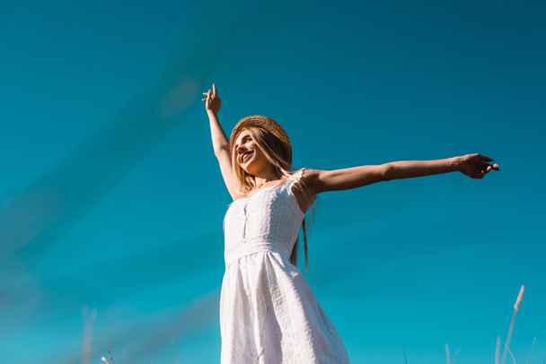 vista de ángulo bajo de la mujer en vestido blanco y sombrero de paja mirando hacia otro lado, mientras que de pie contra el cielo azul con las manos extendidas - Foto, imagen