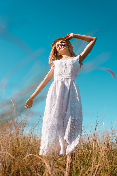 selective focus of woman in white dress touching straw hat while standing with outstretched hand against blue sky - Photo, Image