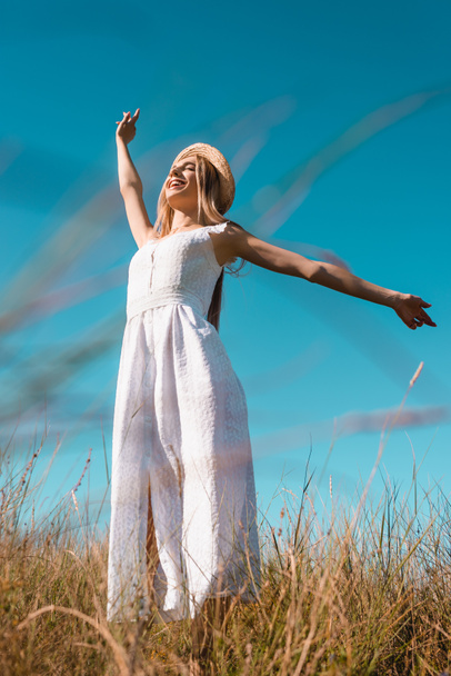 low angle view of stylish woman in white dress standing with outstretched hands and closed eyes against blue sky - Zdjęcie, obraz