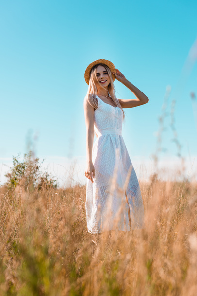 selective focus of young blonde woman in white dress touching straw hat while posing in field against blue sky - Fotoğraf, Görsel