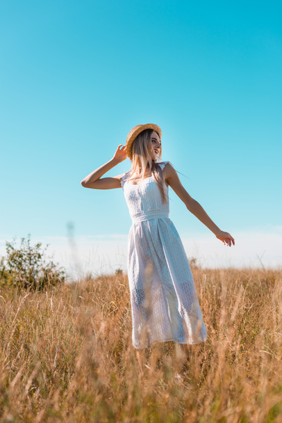 selective focus of blonde woman in white dress and straw hat showing follow me gesture in grassy meadow - Valokuva, kuva