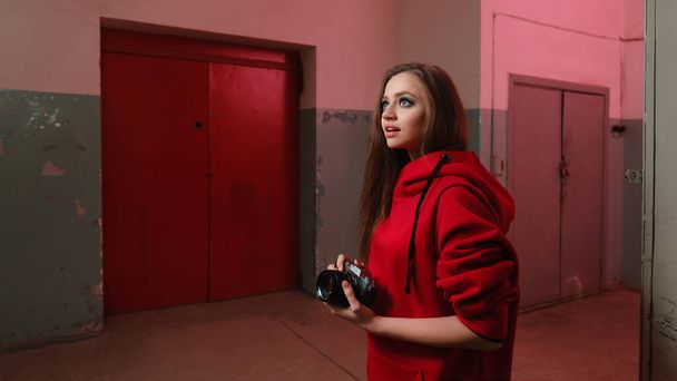 A beautiful girl of 20-25 years old in a red sweatshirt holds a mirrorless camera in an underground room.  - Foto, Imagen