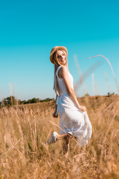 Beyaz elbiseli ve saman şapkalı kadının çayırda poz verirken tek bacağının üzerinde durduğu seçici odak noktası. - Fotoğraf, Görsel