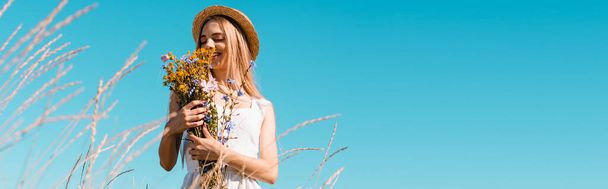 selective focus of young woman in straw hat holding bouquet of wildflowers against blue sky, panoramic concept - Fotó, kép