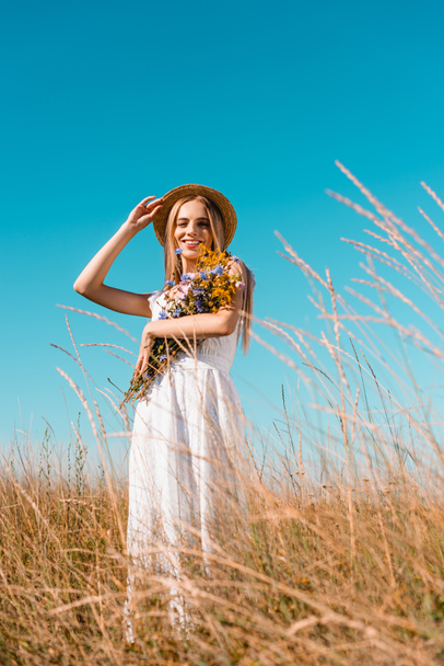 selective focus of young blonde woman in white dress holding wildflowers and touching straw hat while looking at camera - 写真・画像