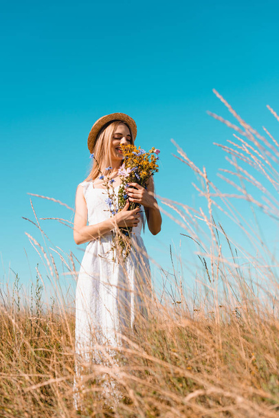 selective focus of blonde woman in straw hat and white dress holding bouquet of wildflowers against blue sky - Foto, imagen
