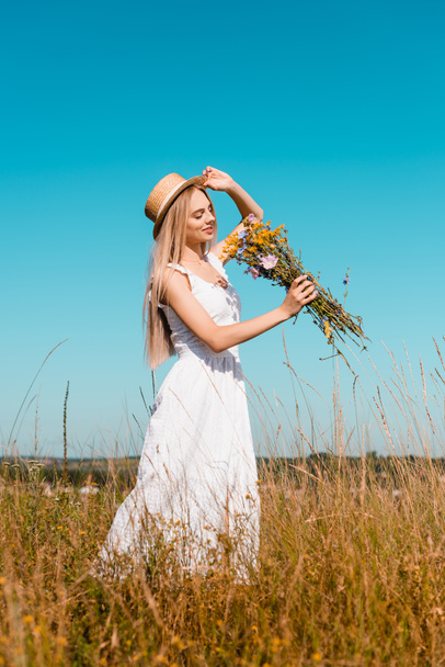 selective focus of sensual woman in white dress touching straw hat while holding wildflowers against blue sky - Zdjęcie, obraz