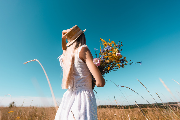 selective focus of young woman in white dress touching straw hat while holding bouquet of wildflowers against blue sky - Photo, Image