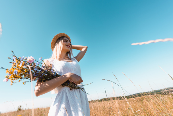 Hochwinkelaufnahme einer jungen Frau mit Wildblumen, die Strohhut berührt, während sie gegen den blauen Himmel wegschaut - Foto, Bild