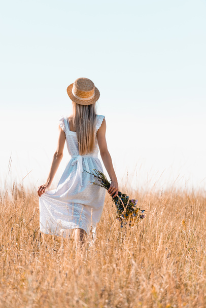 vista posterior de la mujer en sombrero de paja tocando vestido blanco mientras camina en prado herboso con ramo de flores silvestres - Foto, Imagen