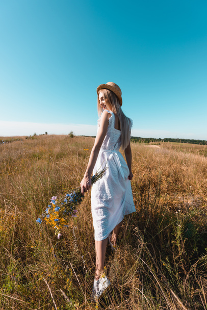 Beyaz elbiseli ve hasır şapkalı genç bir kadın çimenli çayırlarda kır çiçekleriyle yürüyor. - Fotoğraf, Görsel