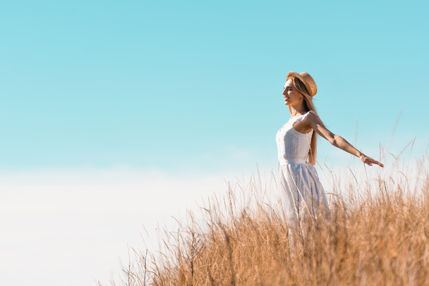 blonde woman in straw hat and white dress standing with outstretched hands on hill against blue sky - Foto, Imagen