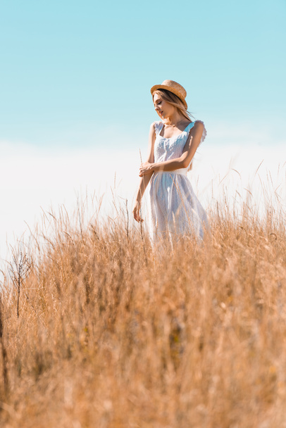 selective focus of young woman in white dress and straw hat looking away while standing on grassy hill - Photo, image
