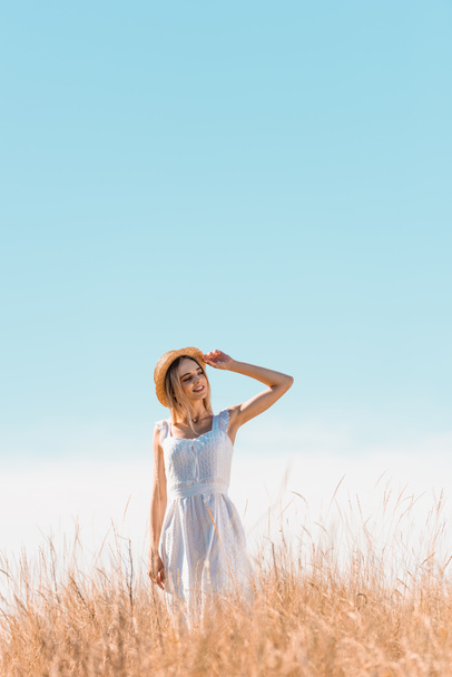 jovem mulher de vestido branco em pé na colina gramada, tocando chapéu de palha e olhando para longe contra o céu azul - Foto, Imagem