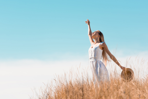 selective focus of blonde woman in white dress holding straw hat while standing with raised hand on grassy hill - Photo, Image