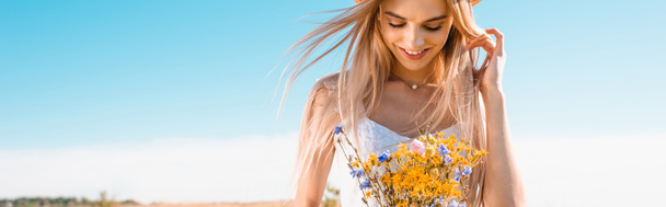 panoramic crop of sensual blonde woman holding bouquet of wildflowers against blue sky - Photo, Image