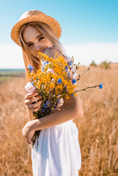 foyer sélectif de la femme blonde en chapeau de paille tenant des fleurs sauvages tout en regardant la caméra dans prairie herbeuse - Photo, image
