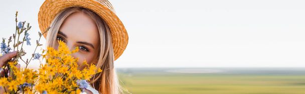 panoramic shot of blonde woman in straw hat holding wildflowers and looking at camera against clear sky and green field - Foto, afbeelding