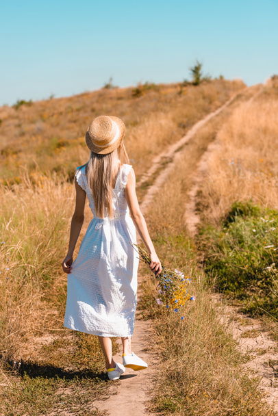 back view of young woman in white dress and straw hat holding wildflowers while walking on road in meadow - Photo, image