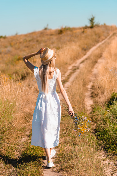 back view of young woman in white dress touching straw hat while walking on road in field with bouquet of wildflowers - Foto, Imagem