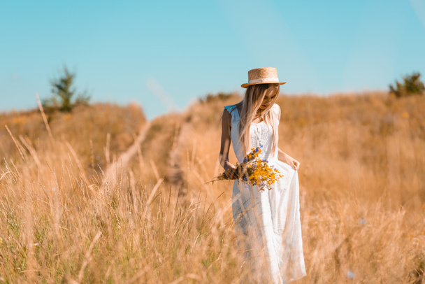 młoda blondynka kobieta w słomkowym kapeluszu dotykając białej sukienki stojąc na polu z bukietem dzikich kwiatów - Zdjęcie, obraz