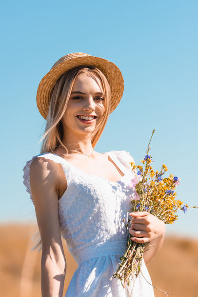 young woman in white dress and straw hat holding bouquet of wildflowers and looking at camera against blue sky - Фото, изображение