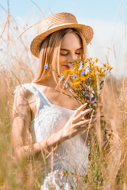 selective focus of stylish woman in straw hat smelling bouquet of wildflowers in grassy meadow - Fotoğraf, Görsel