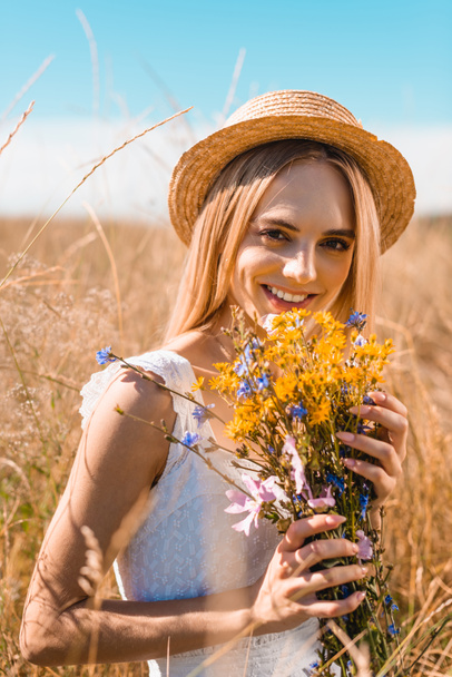 selective focus of blonde woman in straw hat looking at camera while holding wildflowers in grassy meadow - Foto, Imagem