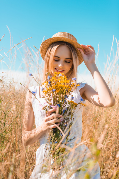 selective focus of blonde woman in white dress touching straw hat while holding wildflowers in grassy field - Fotoğraf, Görsel