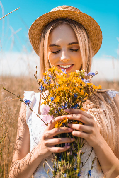 joven rubia en sombrero de paja sosteniendo ramo de flores silvestres en prado herboso - Foto, imagen