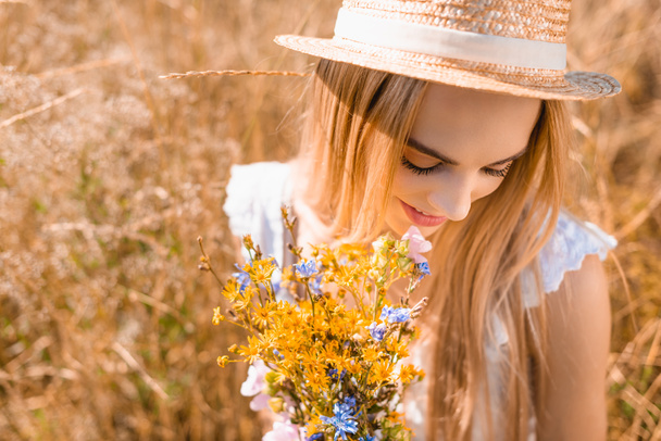 high angle view of sensual blonde woman in straw hat holding bouquet of wildflowers in grassy meadow - Photo, image