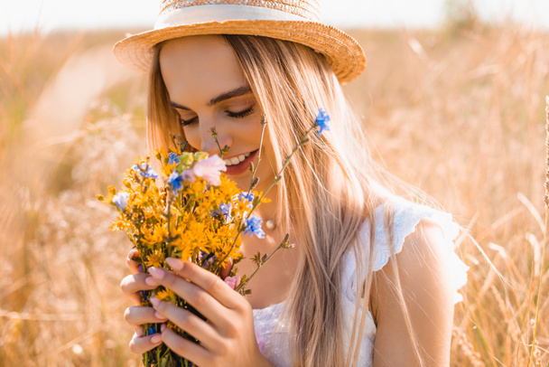 selective focus of blonde woman in straw hat smelling bouquet of wildflowers in grassy meadow - Photo, Image