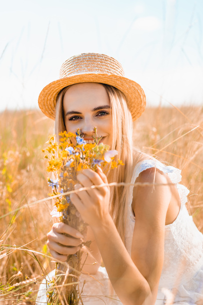 selective focus of blonde woman in straw hat looking at camera while holding wildflowers in grassy meadow - Valokuva, kuva