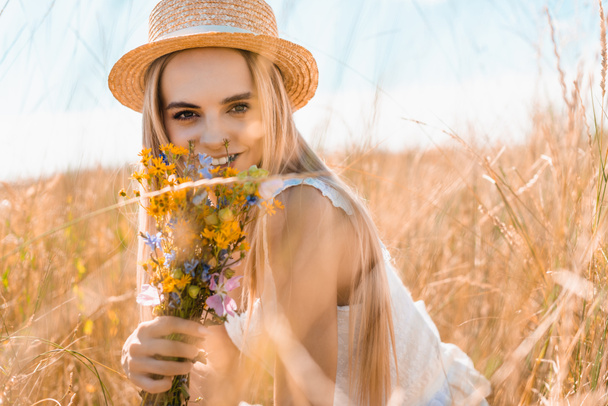 foco seletivo da jovem mulher em chapéu de palha segurando flores silvestres e olhando para a câmera no prado gramado - Foto, Imagem