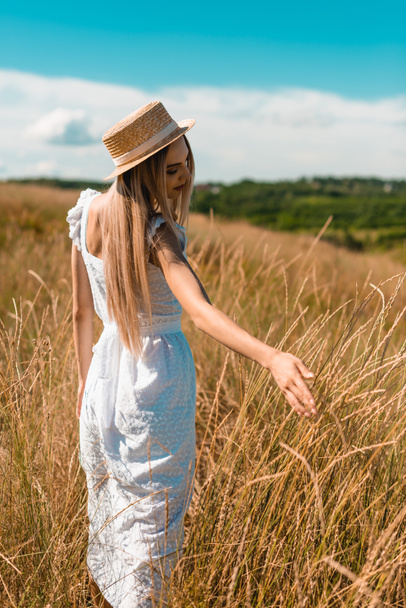 joven mujer rubia en vestido blanco y sombrero de paja tocando espiguillas mientras está de pie en el campo - Foto, imagen