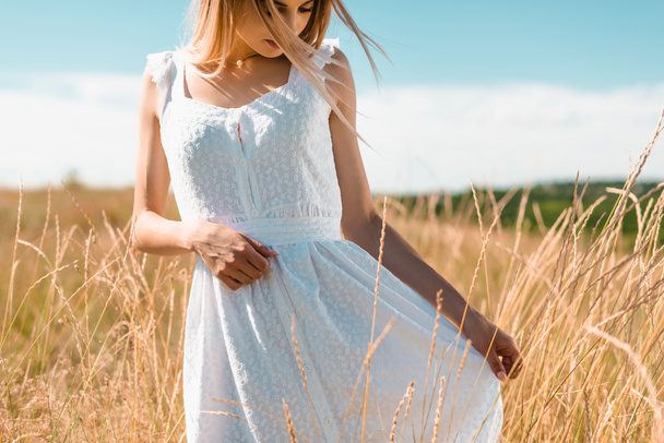jeune femme touchant robe blanche tout en se tenant debout dans un champ herbeux - Photo, image