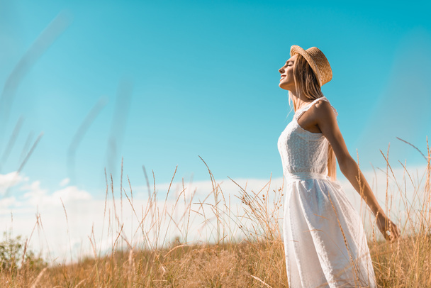 selective focus of stylish woman in white dress and straw hat standing with outstretched hands and closed eyes in grassland - 写真・画像