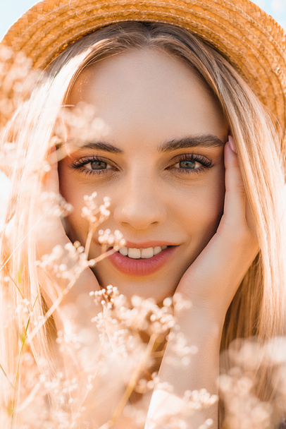 portrait of sensual blonde woman in straw hat looking at camera and touching face near wildflowers - Photo, Image