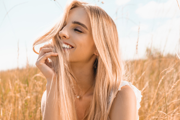 sensual blonde woman touching hair and looking away near grass in field - Foto, afbeelding