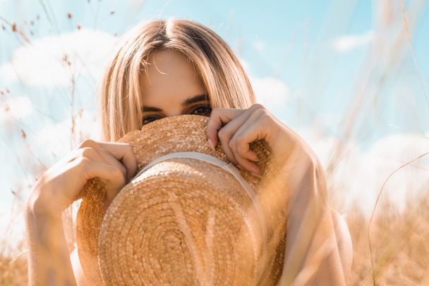 selective focus of blonde woman obscuring face with straw hat while looking at camera against blue sky - Fotografie, Obrázek